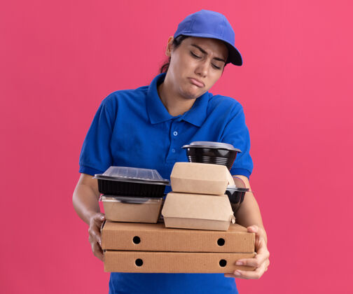 表情悲伤的年轻送货女孩 穿着制服 戴着帽子 把食物容器放在隔离在粉红色墙上的比萨饼盒上帽子姿势食物