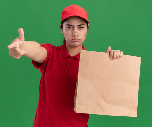 点自信的年轻送货女孩穿着制服 戴着帽子拿着纸食品包装点对着隔离在绿色墙上的摄像机食物衣服包装