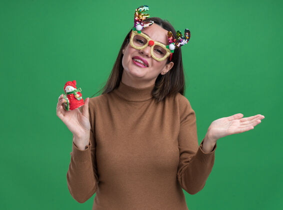 微笑微笑的年轻漂亮女孩穿着棕色毛衣 戴着圣诞眼镜 手里拿着圣诞玩具 手被隔离在绿色的墙上蔓延抱着年轻