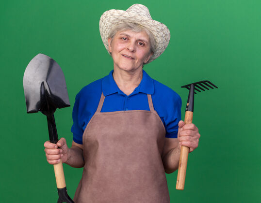 绿色自信的上了年纪的女园丁戴着园艺帽拿着铲子和耙子黑桃耙花园