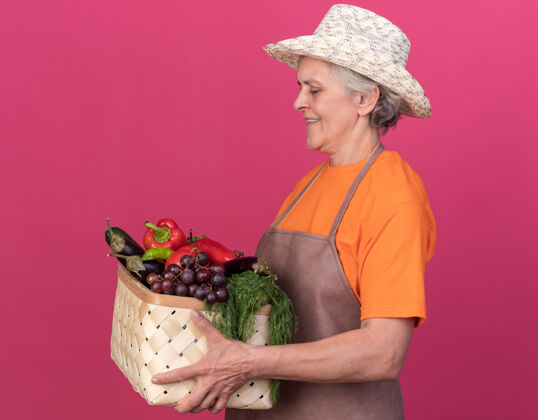 抱着带着园艺帽的上了年纪的女园丁微笑着站在一旁 手里拿着菜篮 看着菜篮园艺侧着帽子