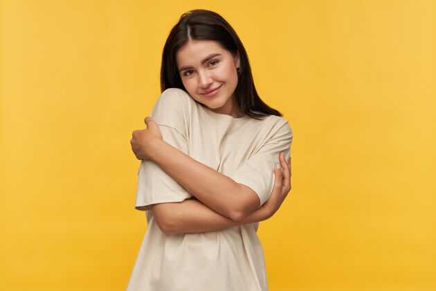 姿势可爱迷人的黑发年轻女子的肖像 穿着白色T恤 站在黄色的墙上 双手相拥女孩假装女人