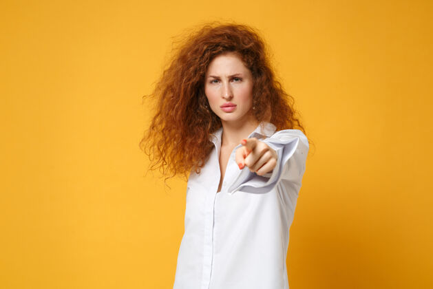站立不满意的年轻红发女女孩穿着休闲白衬衫在黄橙色的墙上摆出孤立的姿势指向食指人物