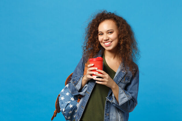 杯子年轻的非洲裔美国女孩十几岁的学生在牛仔服装 背包举行纸杯隔离在蓝色的墙壁上女性红色站