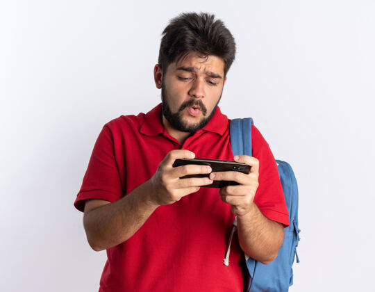玩年轻的留着胡子的学生 穿着红色马球衫 背着背包 用智能手机玩游戏 站在白色的墙上 看起来很担心年轻马球担心