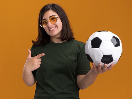 年轻微笑着的年轻漂亮的高加索女孩戴着太阳眼镜拿着并指着球抱着漂亮指着