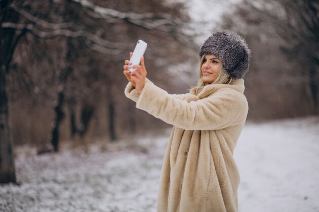 户外穿着冬衣的女人走在满是雪的公园里打电话寒冷树美丽