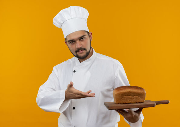 指向身着厨师制服的自信的年轻男厨师手拿着并指着橘色墙上挂着面包的砧板男性手烹饪