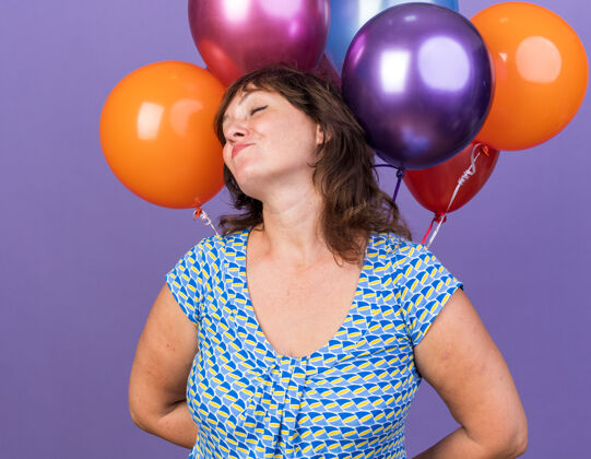 站中年妇女手里拿着一束五颜六色的气球 喜气洋洋地笑着站在紫色的墙上欢庆生日派对欢呼庆祝五颜六色