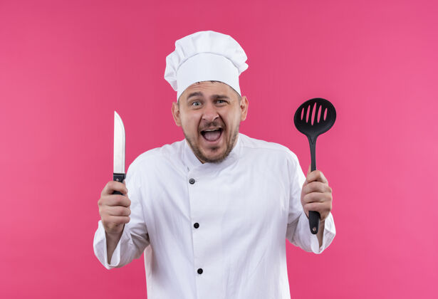 印象深刻年轻帅气的厨师穿着厨师制服 手里拿着开槽汤匙和刀 隔离在粉红色的墙上手持厨师帅气