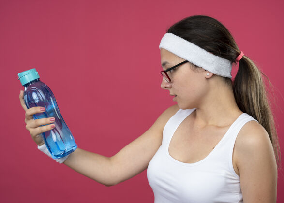 复制自信的年轻运动女孩戴着眼镜戴着头带和腕带拿着和看着水瓶持有头带女孩