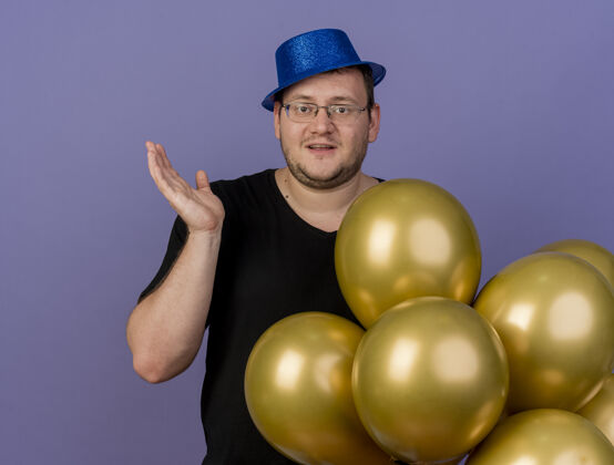 复制高兴的成年斯拉夫人戴着眼镜 戴着蓝色派对帽 举起手站在氦气球旁边请站立聚会