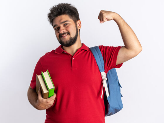 举起来年轻的留着胡子的学生 穿着红色马球衫 背着书包 拿着笔记本 开心而积极地举起拳头微笑着站着背包抱着小伙子