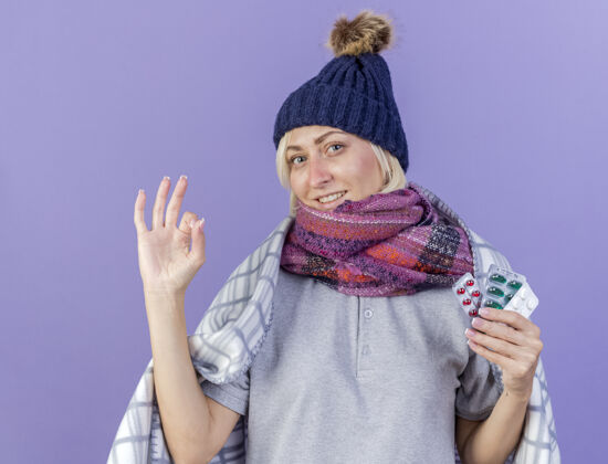 手势微笑着的年轻金发病斯拉夫妇女戴着冬季帽子和围巾裹着格子手势ok手势围巾包装医疗