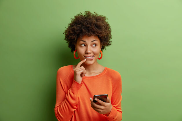 姿势长得漂亮的卷发年轻女子手持现代智能手机使用新应用程序穿着休闲橙色毛衣隔离在绿色墙壁上思考一些令人愉快的事情秋天手机跳投