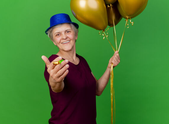 老人微笑着戴着派对帽的老妇人手持氦气球 吹着绿色的口哨哨子氦派对