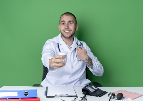 朝向面带微笑的年轻男性医生穿着医用长袍 听诊器坐在办公桌旁 工作工具将医用烧杯向前伸出 手里拿着隔离在绿色墙上的一包胶囊感觉工具医疗