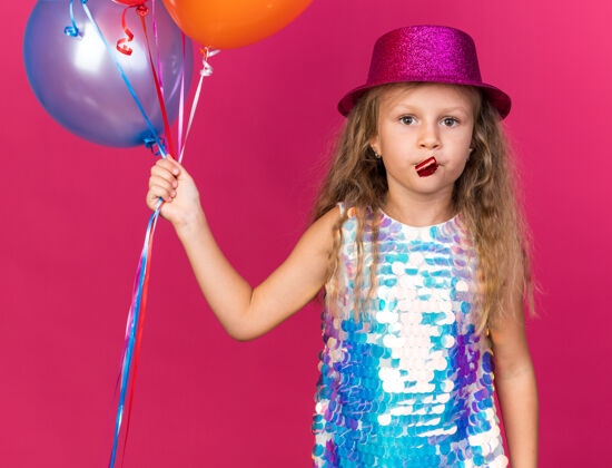 自信自信的金发小女孩 戴着紫色派对帽 手里拿着氦气球 吹着派对哨子 隔离在粉色的墙上 留着复制空间小气球派对