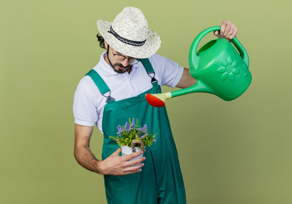 壶年轻的留着胡须的园丁 穿着连体衣 戴着帽子 手里拿着浇水罐 手里拿着盆栽植物 站在淡绿色的墙上浇水市民穿着人