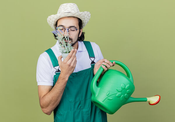 吸入器年轻的留着胡须的园丁身穿连体衣 戴着帽子 手里拿着水罐 手里拿着盆栽植物 站在淡绿色的墙上 散发着宜人的气味人连身衣壶