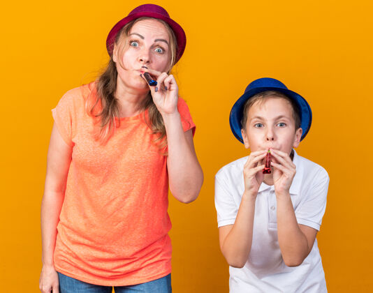 年轻惊讶的年轻斯拉夫男孩戴着蓝色的派对帽 他的母亲戴着紫色的派对帽吹着派对口哨 隔离在橙色的墙上 留着复印空间孤立斯拉夫哨子