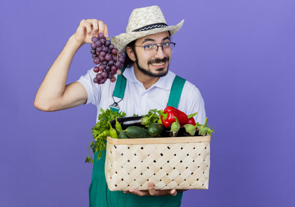 抱着年轻的留着胡须的园丁 穿着连体衣 戴着帽子 手里拿着装满蔬菜的板条箱 蓝色的墙上挂着一束微笑的葡萄人感觉蔬菜
