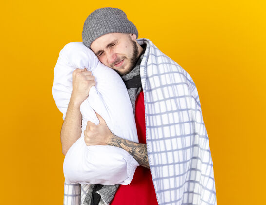 冬天那个戴着冬日帽子 戴着围巾 浑身酸痛的年轻人抱着头 把头放在用格子布包裹的枕头上 隔离在橙色的墙上拥抱穿着表情