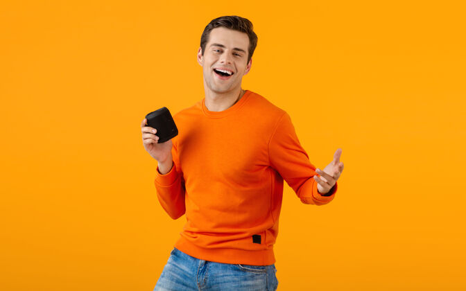 男人穿着橙色毛衣的时髦微笑的年轻人拿着无线扬声器快乐地听音乐享受橙色的乐趣设备时尚休闲