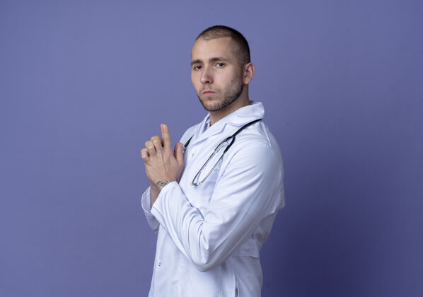 衣服自信的年轻男医生 身穿医用长袍 脖子上戴着听诊器 站在侧视图中 用手做着的手势 看着紫色墙壁上孤立的前方医学手势男性