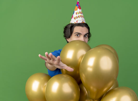 帅气印象深刻的帅哥戴着生日帽站在氦气球伸出双手隔离在绿色的墙上伸展印象人
