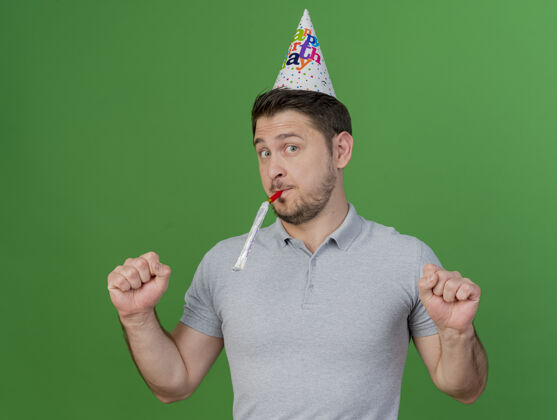生日高兴的年轻人戴着生日帽吹着口哨 表示是的姿态孤立的绿色派对穿帽子小伙子