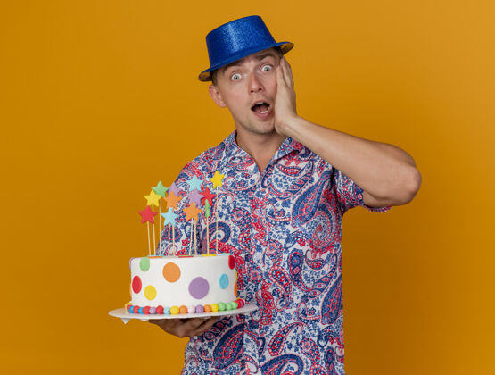抱着惊讶的年轻人戴着蓝色帽子拿着蛋糕 手放在脸颊上孤立的橙色戴着年轻人帽子