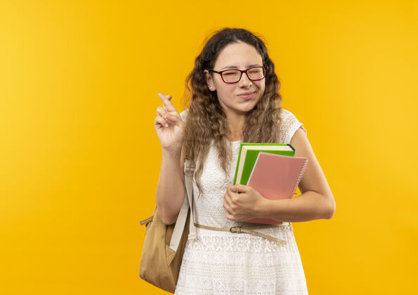眼睛高兴的年轻漂亮的女学生戴着眼镜 背着书包 拿着笔记本 手指交叉 闭着眼睛 隔离在黄色的墙上漂亮眼镜人