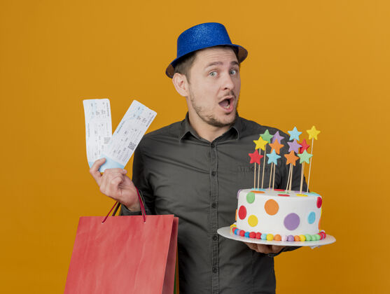 年轻人快乐的年轻人戴着蓝色的帽子 拿着蛋糕和礼品袋 票子孤立在橙色的帽子欢乐蓝色