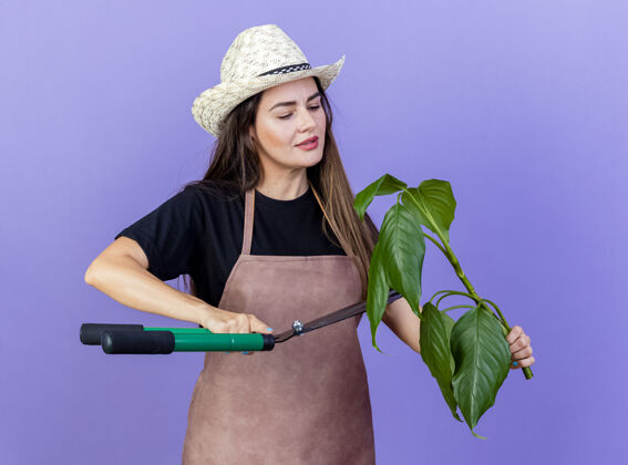 请穿高兴美丽的园丁女孩穿着制服戴着园艺帽拿着修剪植物用剪子隔离在蓝色剪子拿剪
