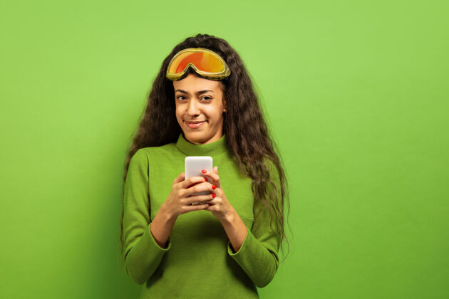 非洲以绿色工作室为背景 黑色皮肤的非洲裔年轻女子的滑雪面具肖像人类情感 面部表情 销售 广告 冬季运动和假期的概念使用智能手机运动女性美国