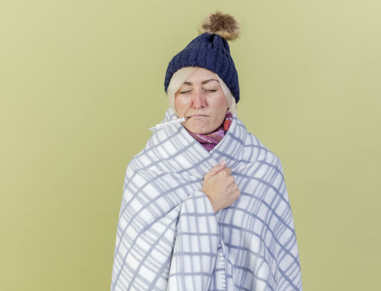 年轻年轻的金发病女人戴着冬天的帽子 围巾裹着格子呢 嘴里叼着温度计 隔离在橄榄绿的墙上包裹人疾病