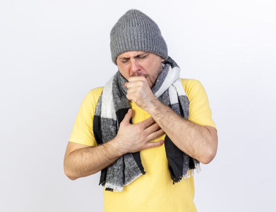 冬天咳嗽的年轻金发病男子戴着冬天的帽子和围巾把手放在胸前隔离在白墙上表情金发胸部
