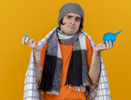 男人困惑的年轻病人戴着冬天的帽子 围巾裹在格子布里 拿着灌肠剂 把双手分开放在橘子上橘子围巾帽子