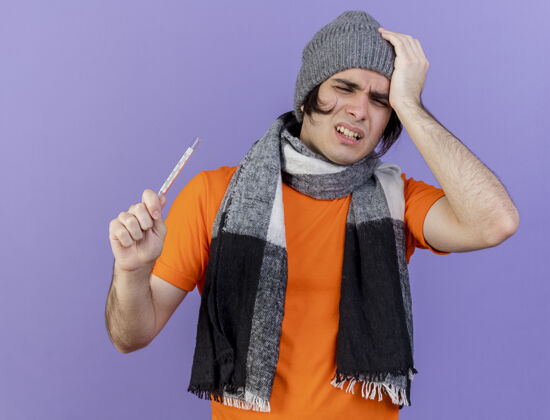 帽子体弱多病的年轻人戴着冬天的帽子 戴着围巾 拿着体温计 把手放在紫色隔离的疼痛的头上男人疼生病
