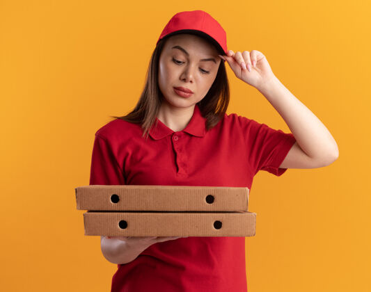 橘子穿着制服的自信漂亮的女送货员把手放在帽子上拿着 看着橙色的披萨盒披萨盒子手