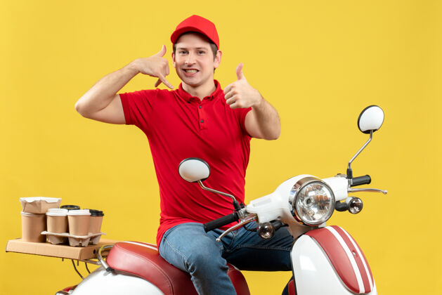 健身黄色墙壁上微笑的年轻人穿着红色上衣 戴着帽子传递订单 打电话给我好的手势电话衬衫摩托车