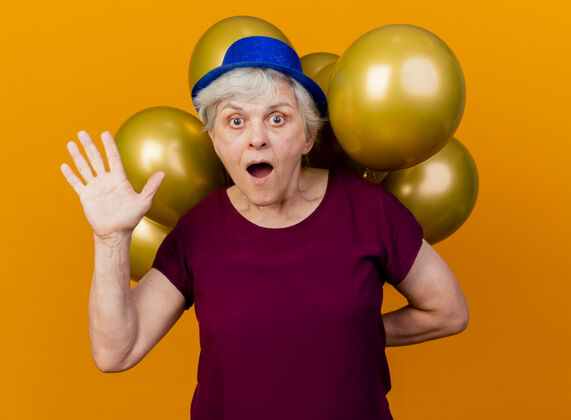 帽子惊讶的戴着派对帽的老妇人举起手来 拿着氦气球 身后是隔离的橘黄色墙壁 有复制空间举老人惊喜