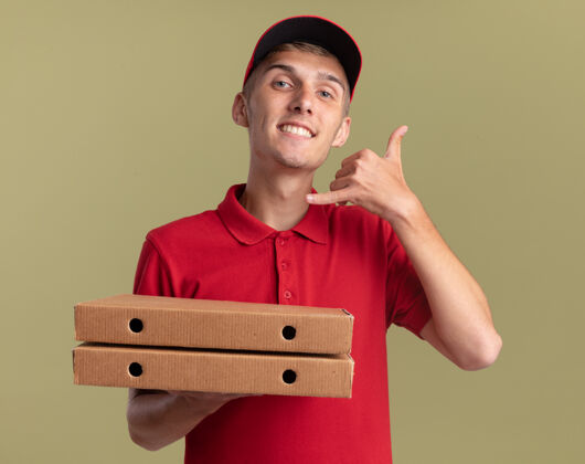 披萨微笑的年轻金发送货男孩拿着比萨饼盒 手势叫我橄榄绿上的标志送货手势微笑