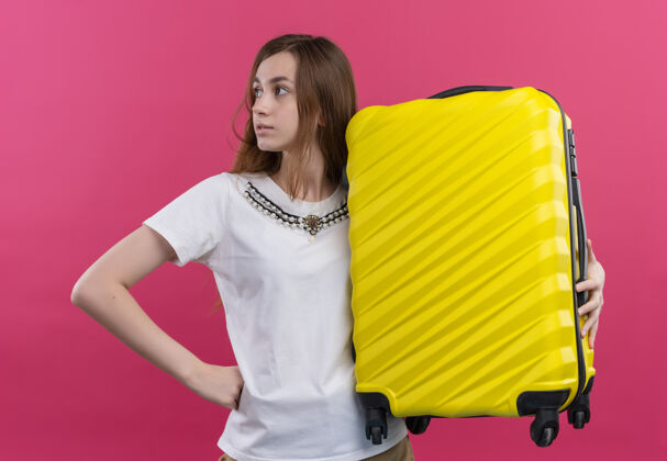 抱着自信的年轻旅行家女孩拿着手提箱在孤立的粉色空间里看着左边旅行者自信女孩