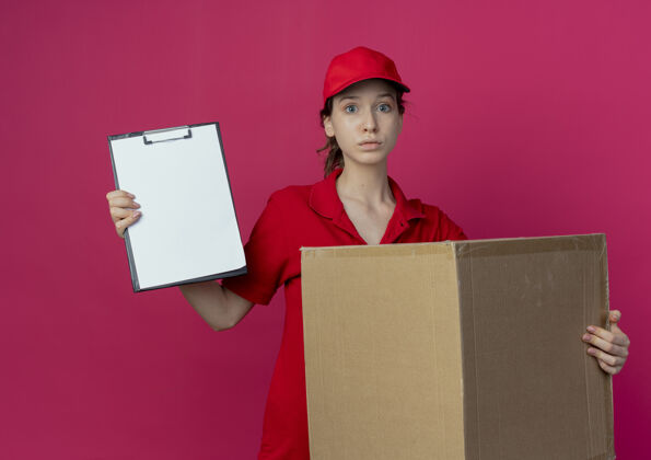 印象令人印象深刻的年轻漂亮的送货女孩在红色制服和帽子举行纸箱和剪贴板孤立的深红色背景年轻漂亮红色