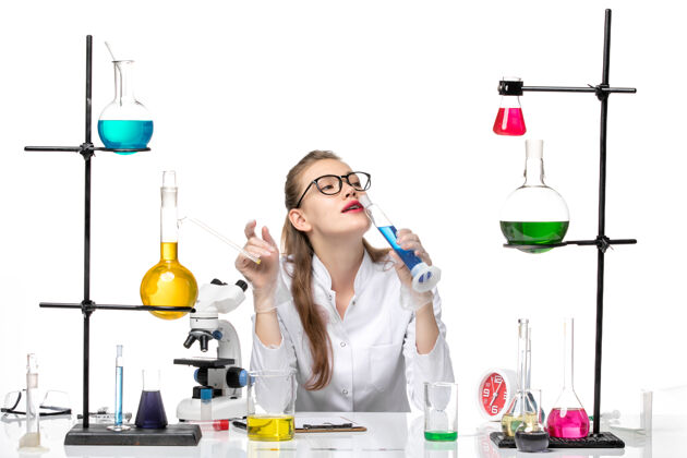 白色正面图白色地板上穿着医疗服的女化学家拿着装有蓝色溶液的长烧瓶化学大流行健康科维德套装烧瓶前面
