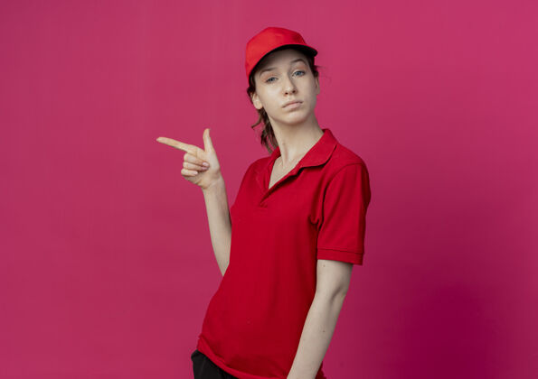 壁板自信的年轻漂亮的送货女孩 穿着红色制服 戴着帽子 指着被隔离在深红色背景上的一边 留着复印空间漂亮女孩背景