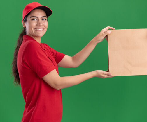 纸面带微笑的年轻送货员 穿着制服 戴着帽子 把纸食品包装交给隔离在绿墙上的客户年轻人帽子食物