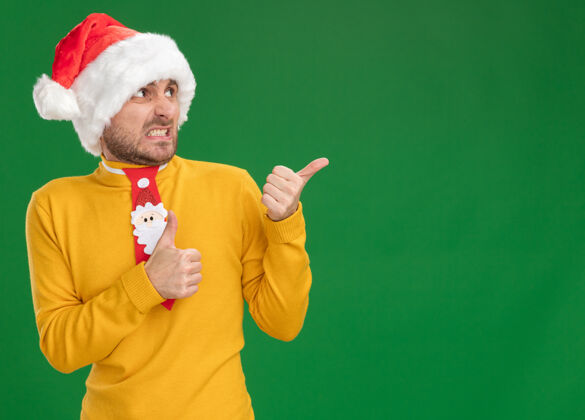 绿色恼怒的年轻白人男子戴着圣诞帽 打着领带 一边看着一边竖起大拇指 孤立地站在绿色背景上人帽子指向